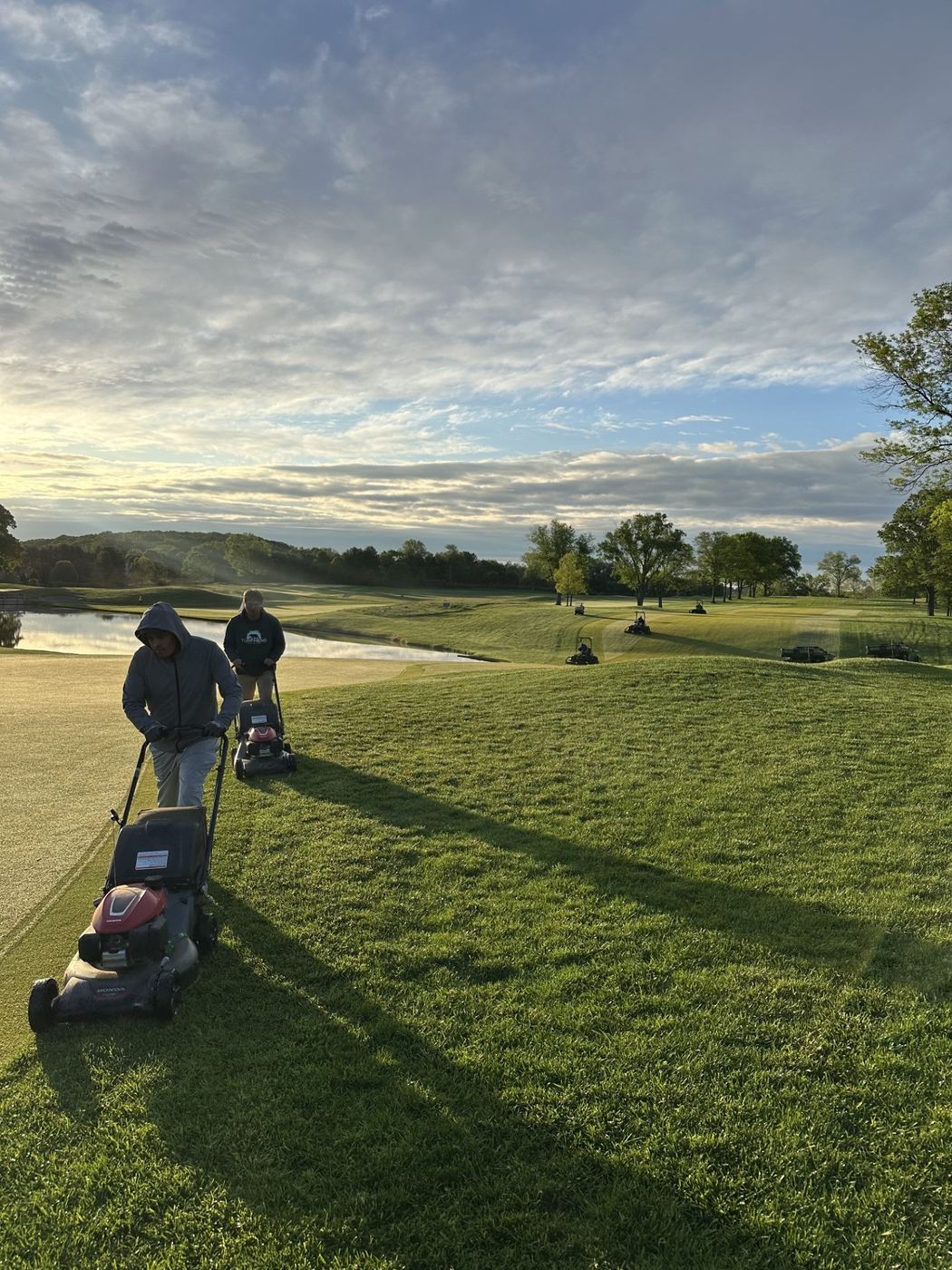 Crew mowing fairways with push mowers at sunrise, with a pond and course carts in the background