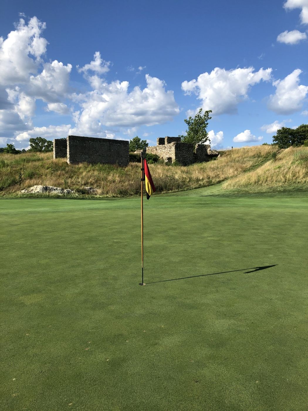 A green at Philadelphia Cricket Club framed by historic stone quarry ruins, with the club's red and yellow flag flying