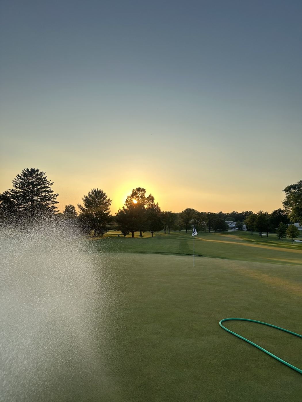 Hose spraying water across a green at sunset, with the flag and fairway visible beyond the mist